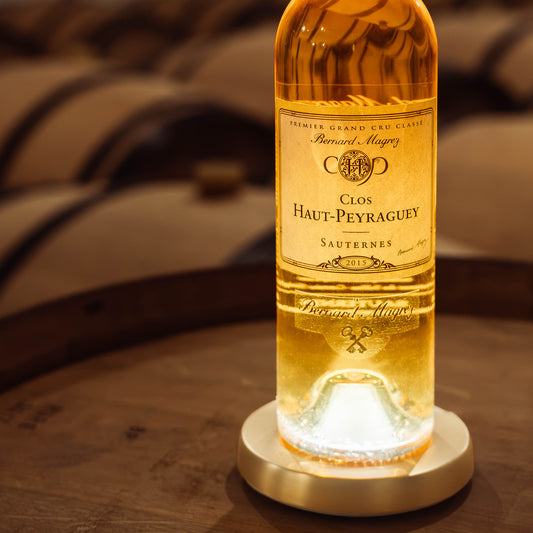 Bouteille de sauternes clos haut Peyraguey posée sur une barrique dans un chai illuminé par dessous de bouteille lumineux yobe light Pro avec coque dorée yobe case
