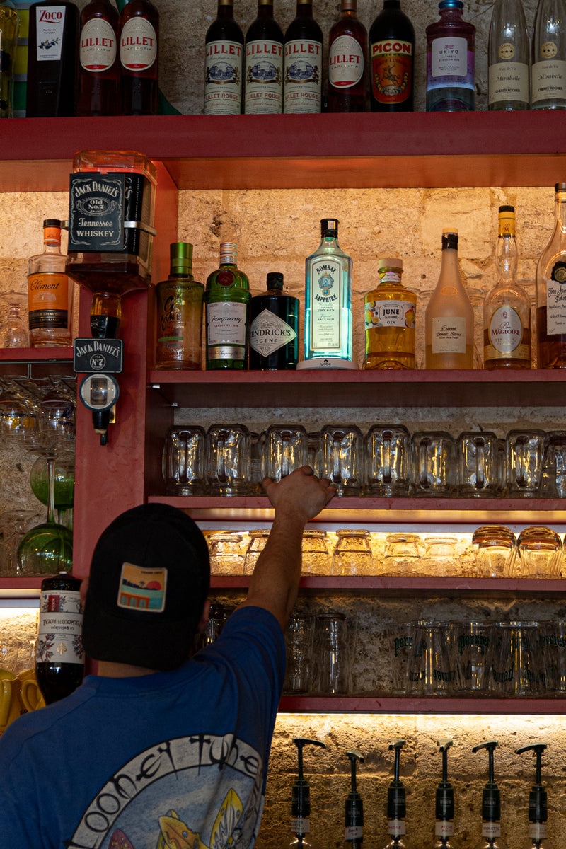 décor de bar avec un homme de dos au premier plan plusieurs bouteilles dont une bleue éclairée par un suppport yobe 