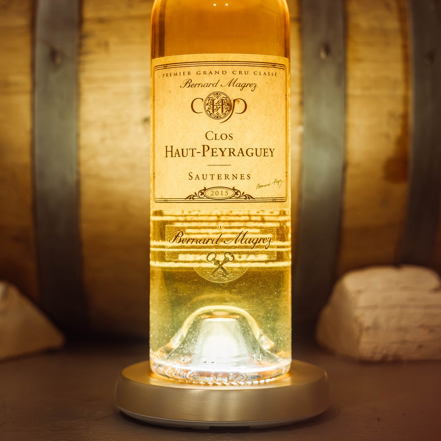 Bouteille de sauternes du clos haut peyraguey illuminée posée sur dessous de bouteille lumineux Yobe light Pro recouvert de la coque yobe case dorée devant une barrique 