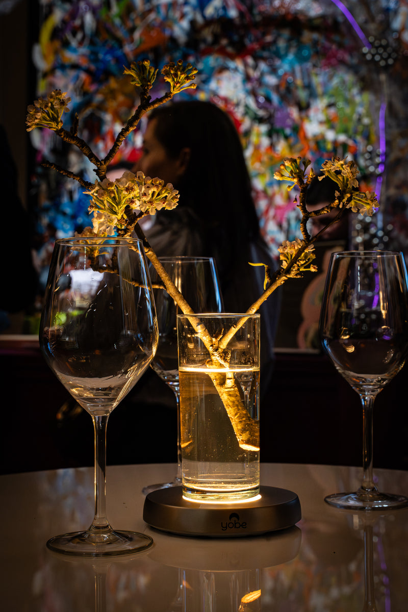 vase avec branches de cerisier posé sur un support lumineux Yobe doré qui l'éclaire d'une lumière chaude. Deux verres à vin sur une table en marbre dans un décor avec des oeuvres d'art  