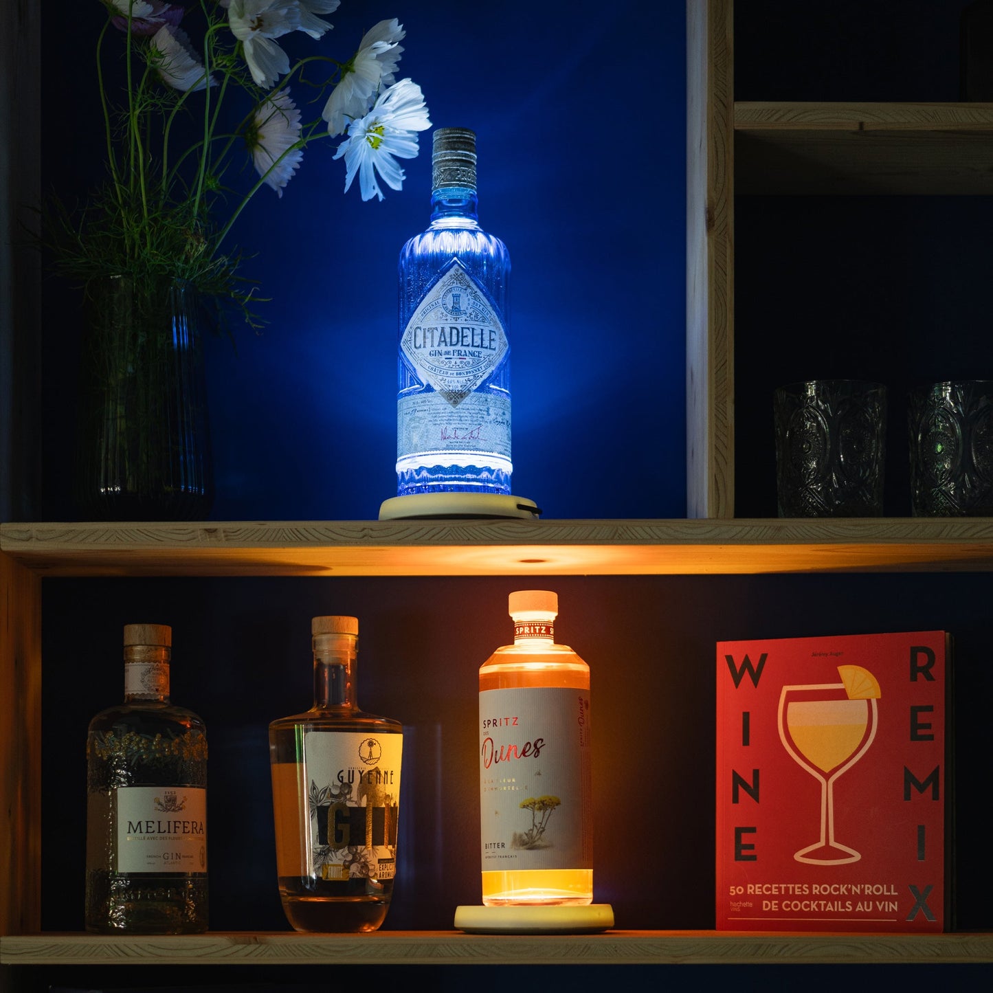 Bouteille lumineuse bleue et bouteille orange posées sur une étagére éclairée avec yobe light dans un décor fleuri et de cocktails