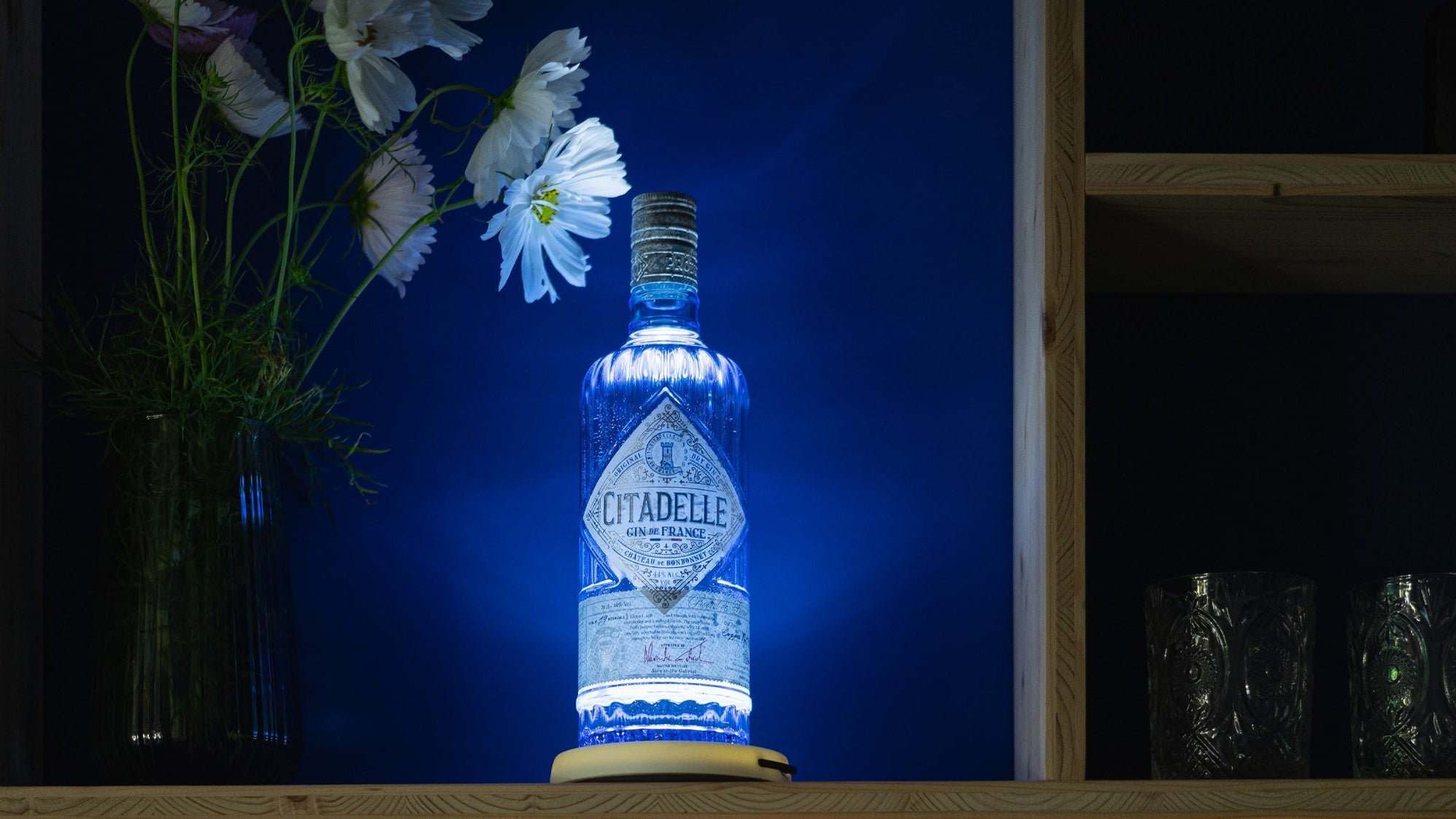 Bouteille lumineuse bleue et bouteille orange posées sur une étagére éclairée avec yobe light dans un décor fleuri et de cocktails